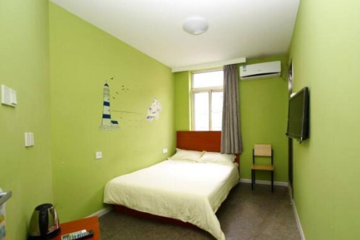 Ein kleines Zimmer mit einem Einzelbett, grünen Wänden, einem Fenster mit Vorhängen, einem Wand-Fernseher und einem Wasserkocher auf dem Boden.