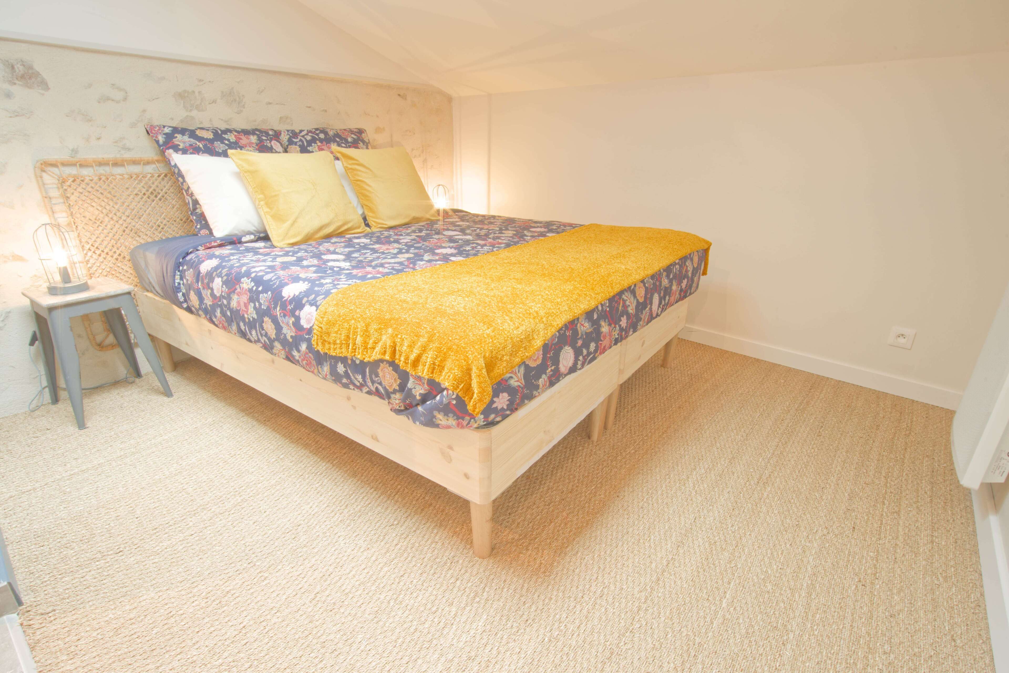 Un dormitorio con una cama de madera, ropa de cama floral, un plaid amarillo, una mesita de noche con una lámpara y una pared con textura.
