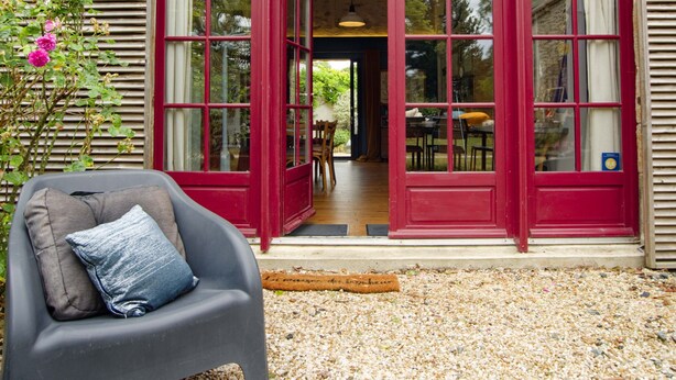 Un edificio con puerta y ventanas rojas, un comedor visible a través de la puerta, y una silla con cojín en el exterior.