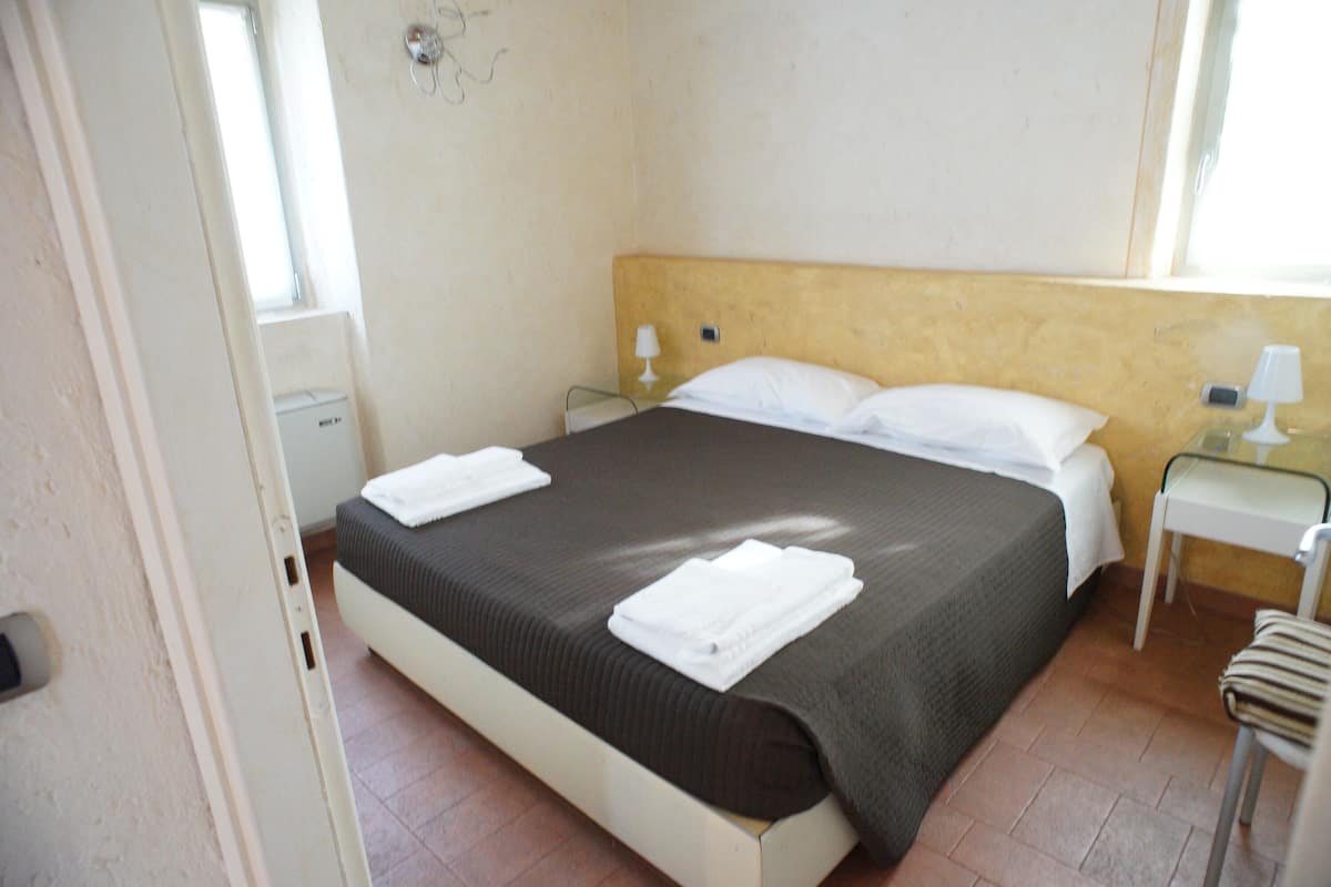 Una cama individual con una colcha marrón y almohadas blancas, una mesita de noche con una lámpara y una silla.