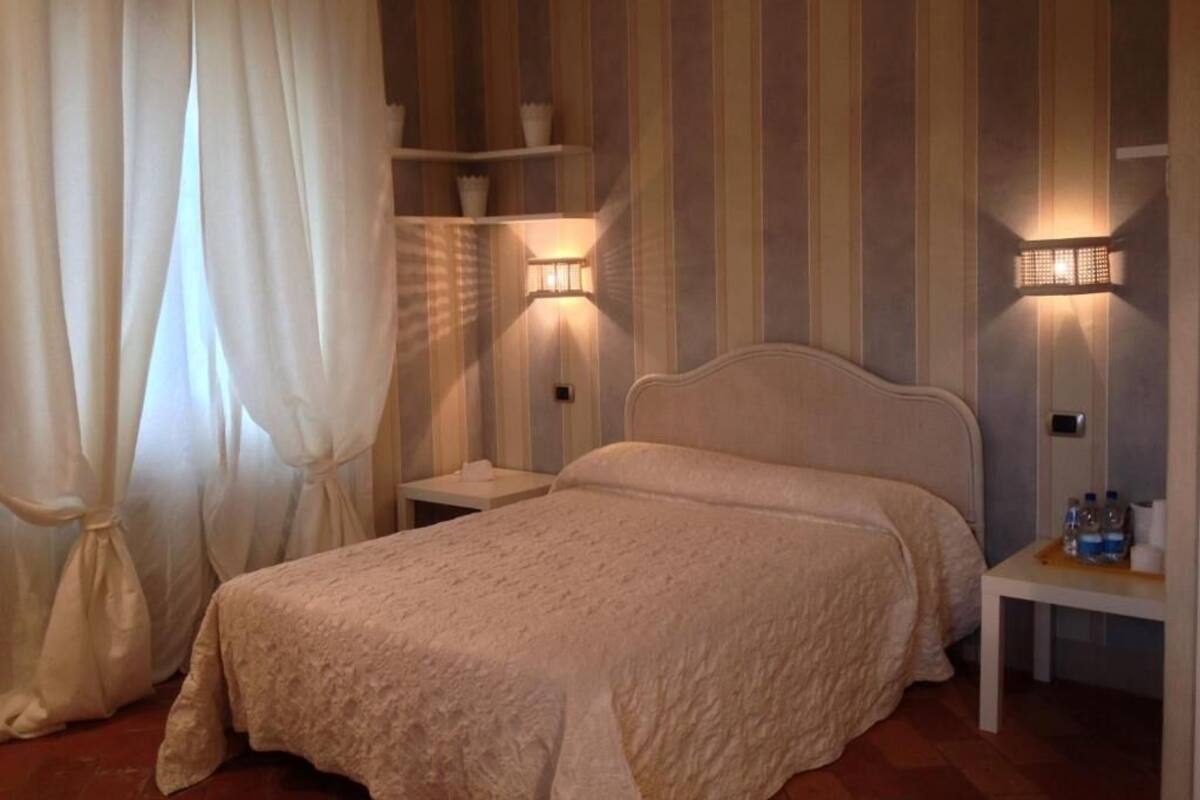 Un dormitorio con cama, cortinas blancas, una mesita de noche con lámpara y estante con objetos decorativos.