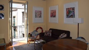 A room with a black leather sofa, a round wooden table, and a chair with a colorful cushion. There are framed artworks on the walls and a floor lamp. - La Baia di Napoli (Naples)