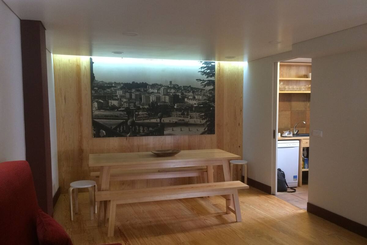 Een woonkamer met een houten bank, een rode bank en een eettafel. Een stadsgezicht schilderij hangt aan de muur.