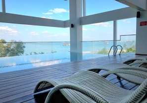 An infinity pool with lounge chairs, large windows, and a view of the sea.
