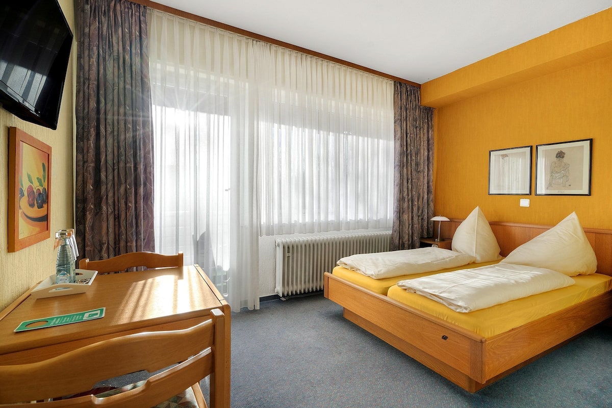 A hotel room with a bed, a desk, and a chair. There is a window with curtains, a radiator, and two framed pictures on the wall.