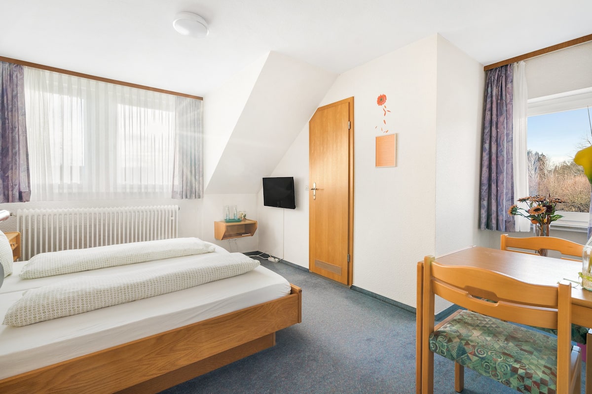 A bedroom with a bed, a wooden desk, a chair, a TV, and a window with curtains.