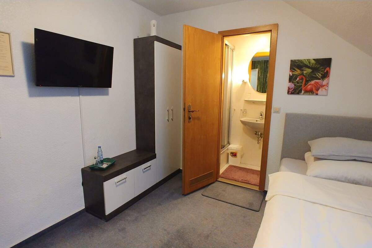 A bedroom with a bed, a TV mounted on the wall, a wardrobe, and a bathroom visible through an open door.
