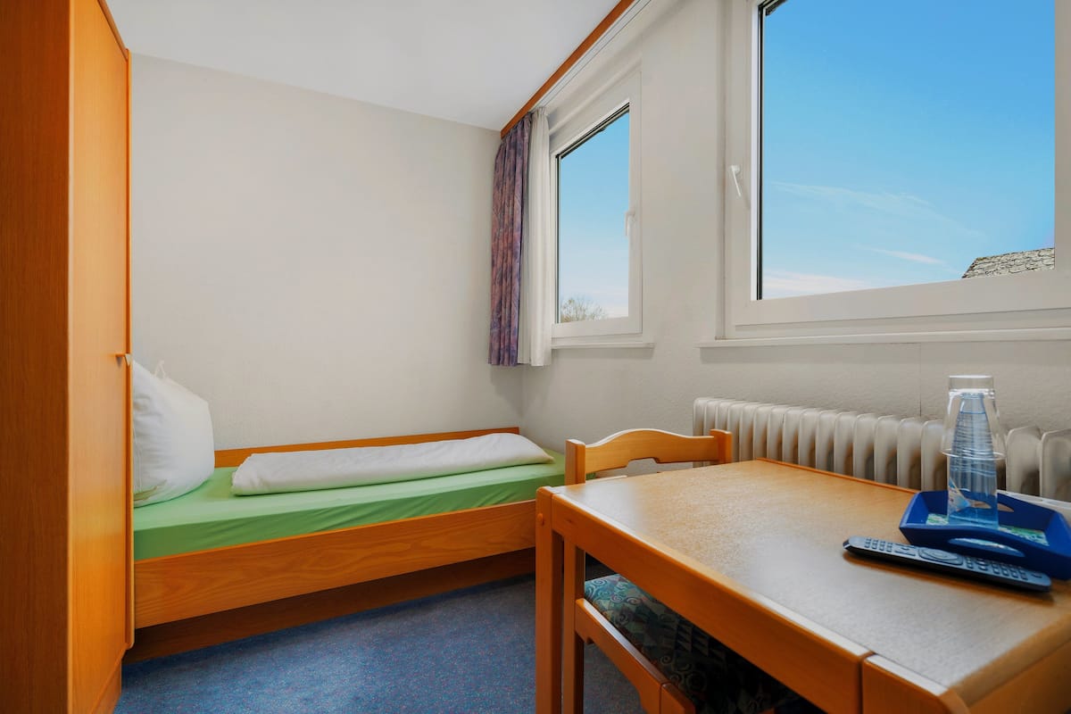 A small room with a single bed, a wooden table, a chair, and a window with a view of the sky.