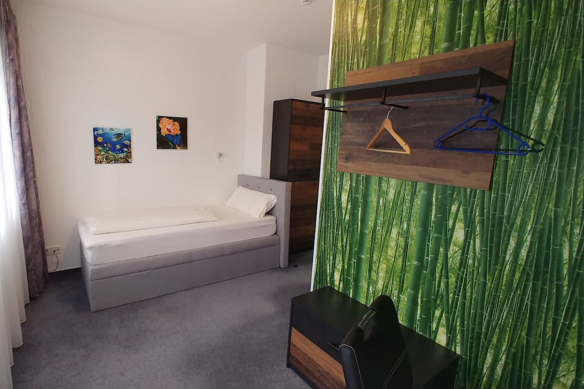 A small, single-bed room with a grey mattress, a wooden headboard, and a green bamboo-patterned wall. There are two framed pictures on the wall, a wooden shelf with a hanger, and a black bag on the floor.