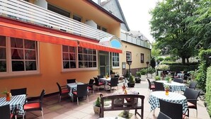 Un espace extérieur aménagé en terrasse avec des tables et des chaises, un bâtiment doté d’un auvent rouge et des plantes en pot.
