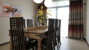 A dining area with a wooden table, chairs, and a vase with yellow flowers. There is a painting on the wall and a window with curtains. - Gurney 4 Plus 1 Bali Style Tranquil Villa (George Town)