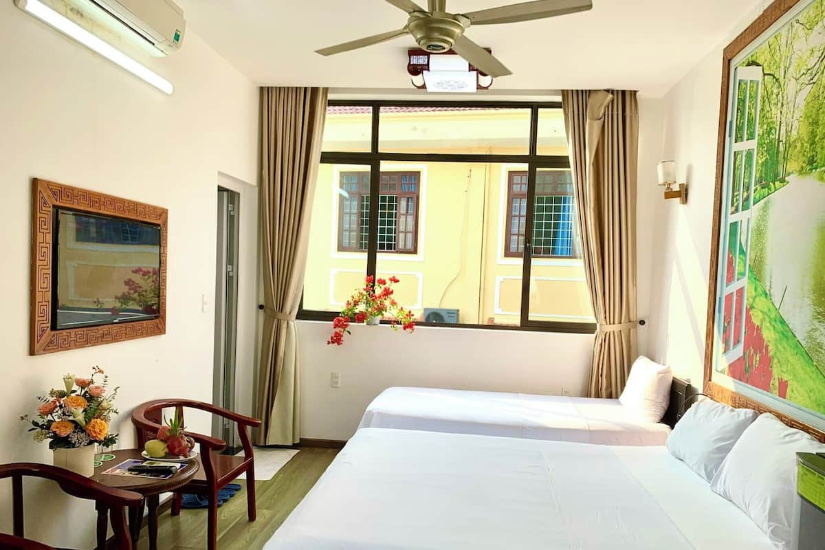 A hotel room with two beds, a desk with a chair, a window with curtains, a ceiling fan, and a painting on the wall.