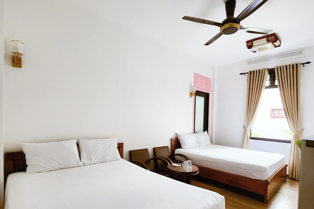 A hotel room with two single beds, a ceiling fan, a small table with a glass, and a window with curtains.