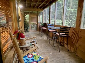 Eine Holzterrasse mit Tisch und Stühlen, einem Teddybären und Blick auf den Wald.