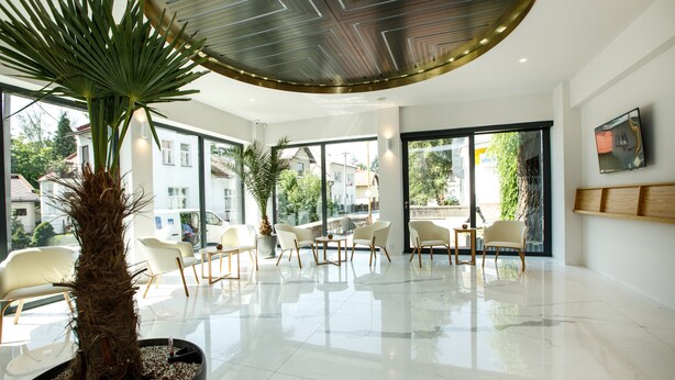 A spacious lobby with a large potted palm tree, modern seating, and large windows offering views of the outside. - Hotel Rajka (Valasske Mezirici)