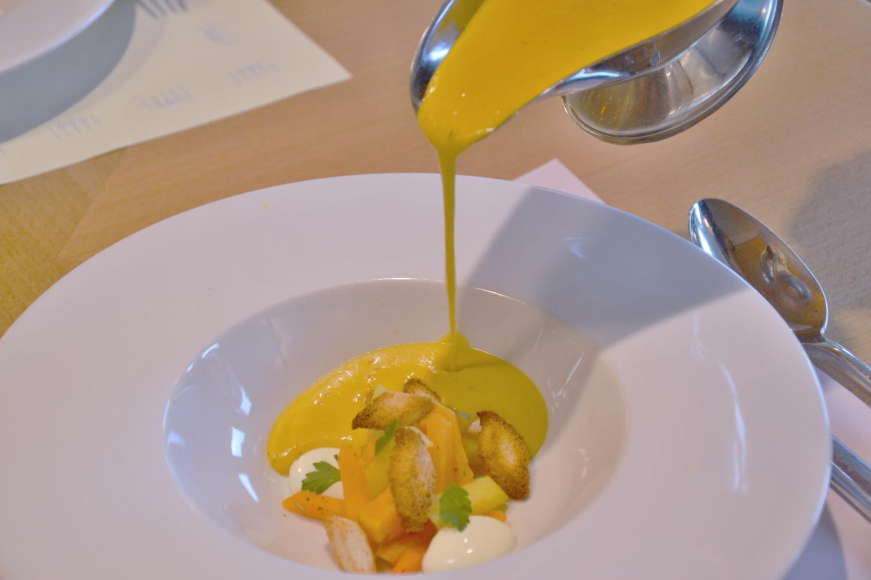 A white plate with a yellow sauce being poured over a dish containing croutons, diced vegetables, and a dollop of cream.