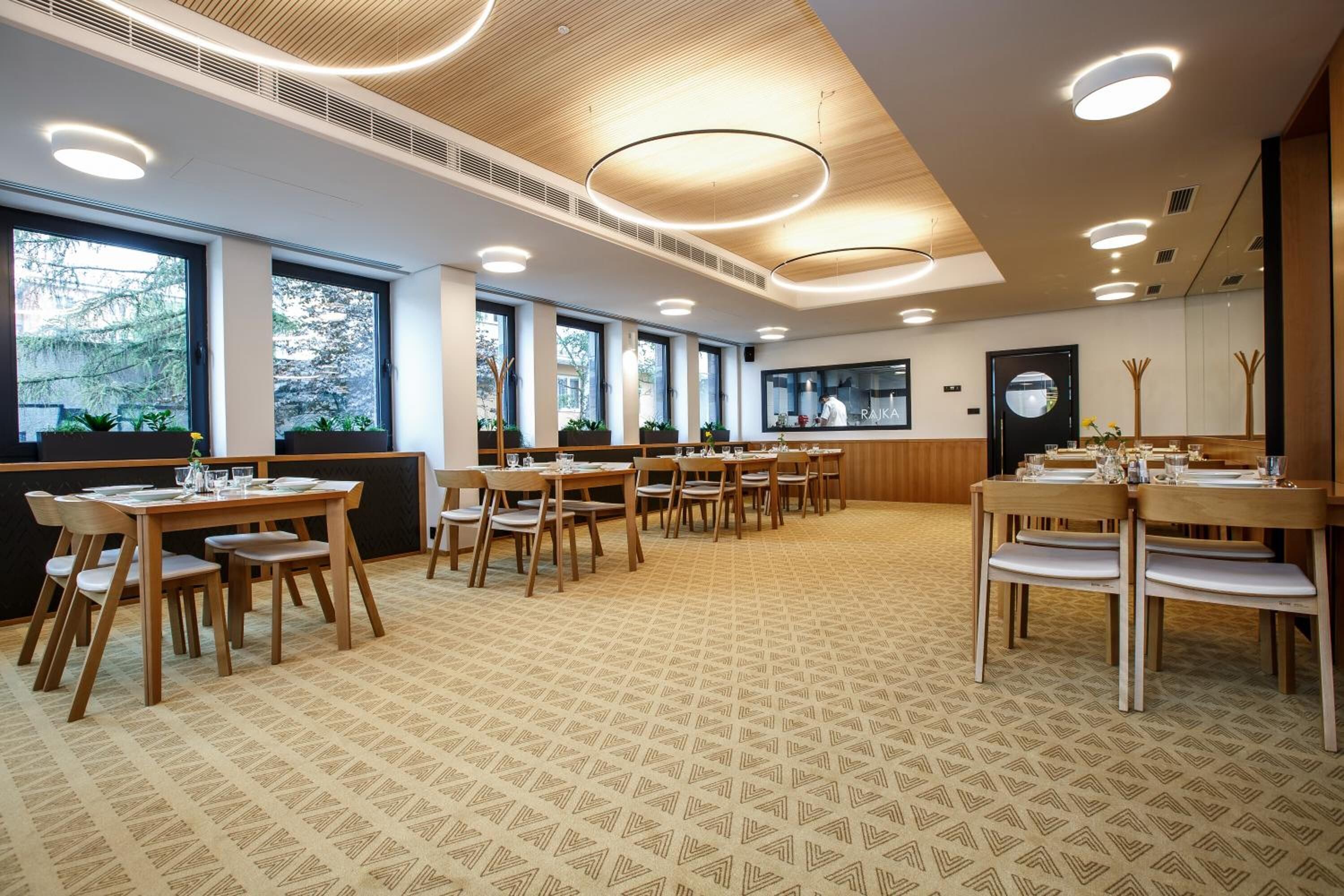 A spacious dining area with multiple tables and chairs, large windows offering views of trees, and a modern ceiling design with circular lighting fixtures.