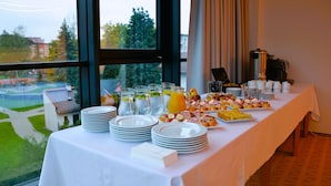 A breakfast buffet table with plates, glasses, and various food items, set against a window with a view of a park and buildings. - Hotel Rajka (Valasske Mezirici)