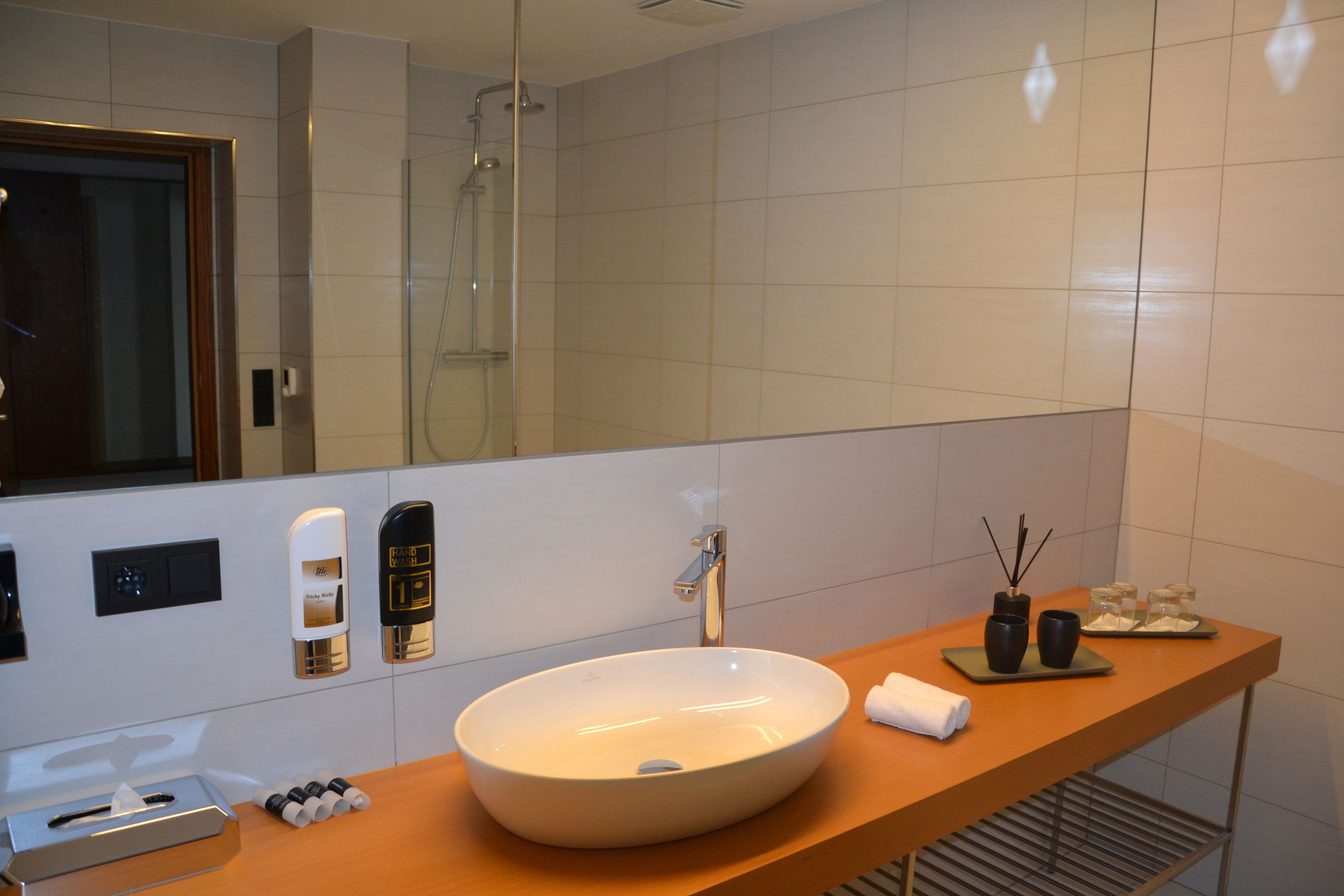 Ein modernes Badezimmer mit einem großen, ovalen Waschbecken, einer Handbrause und einem Spiegel über dem Waschbecken. Auf der Ablage liegen Pflegeprodukte und ein Handtuch hängt an der Wand.