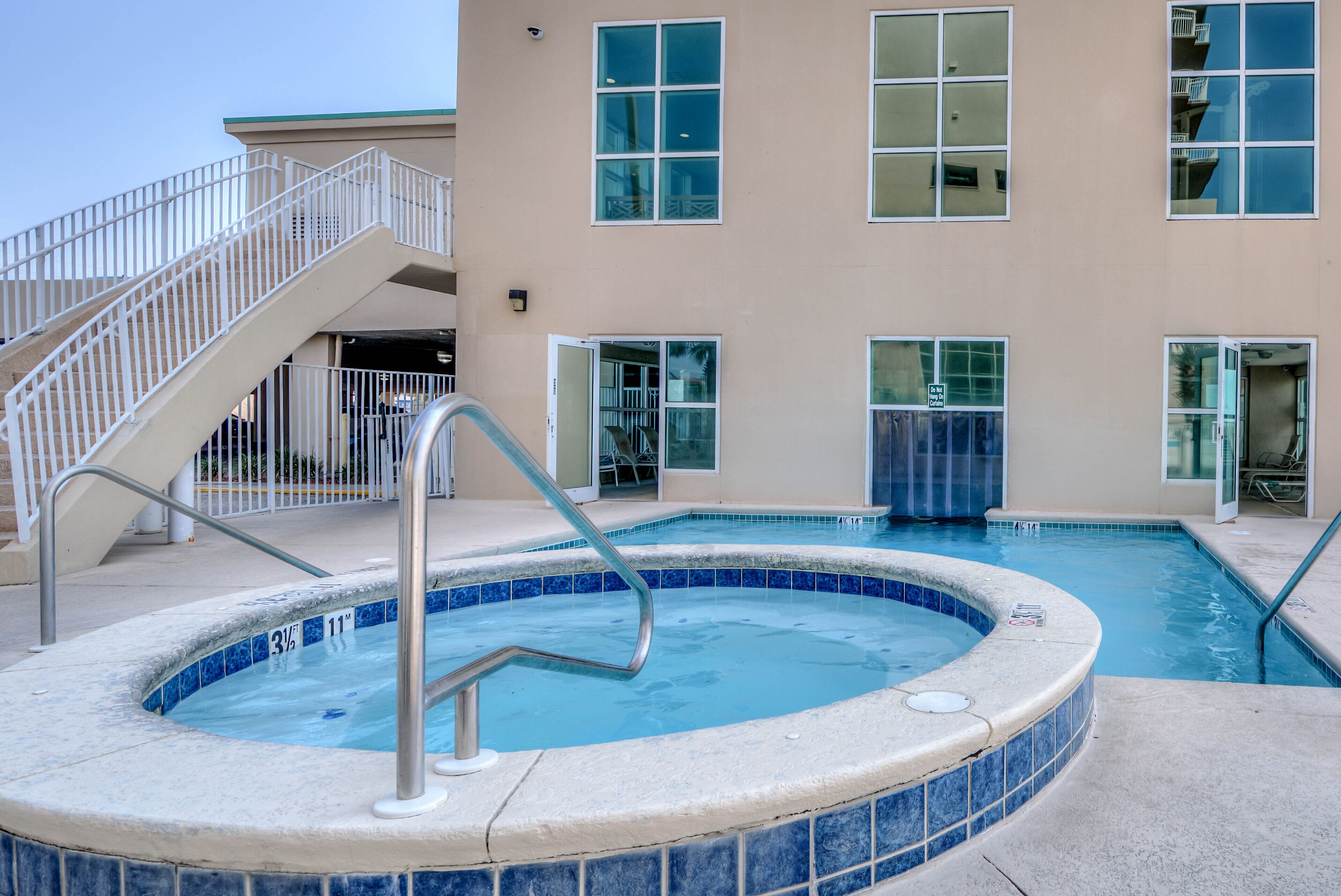 A swimming pool with a tiled edge, a metal handrail, and a stairway leading to a building with large windows.