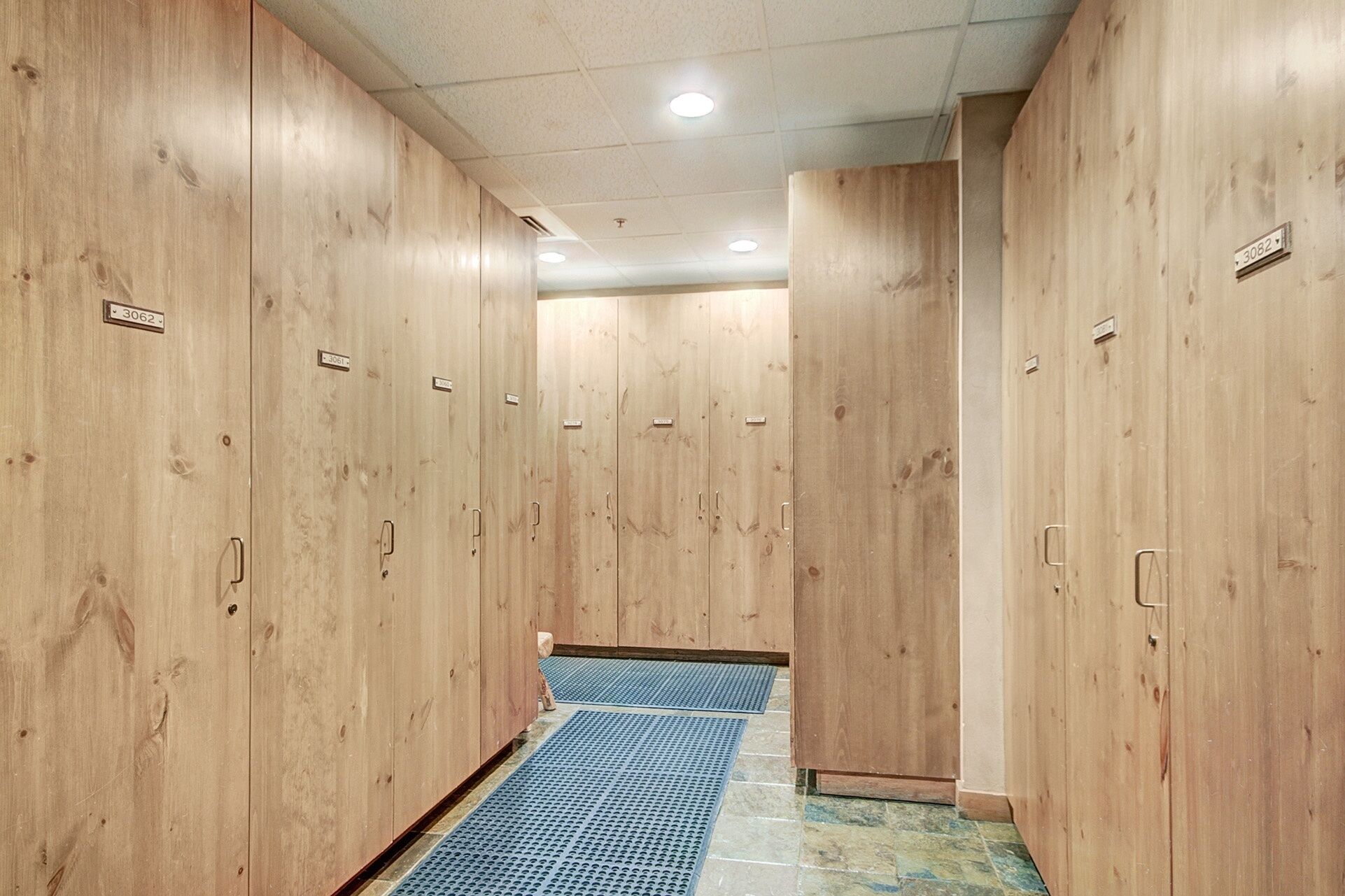 A hallway with wooden lockers, a blue mat at the entrance, and a ceiling with recessed lighting.