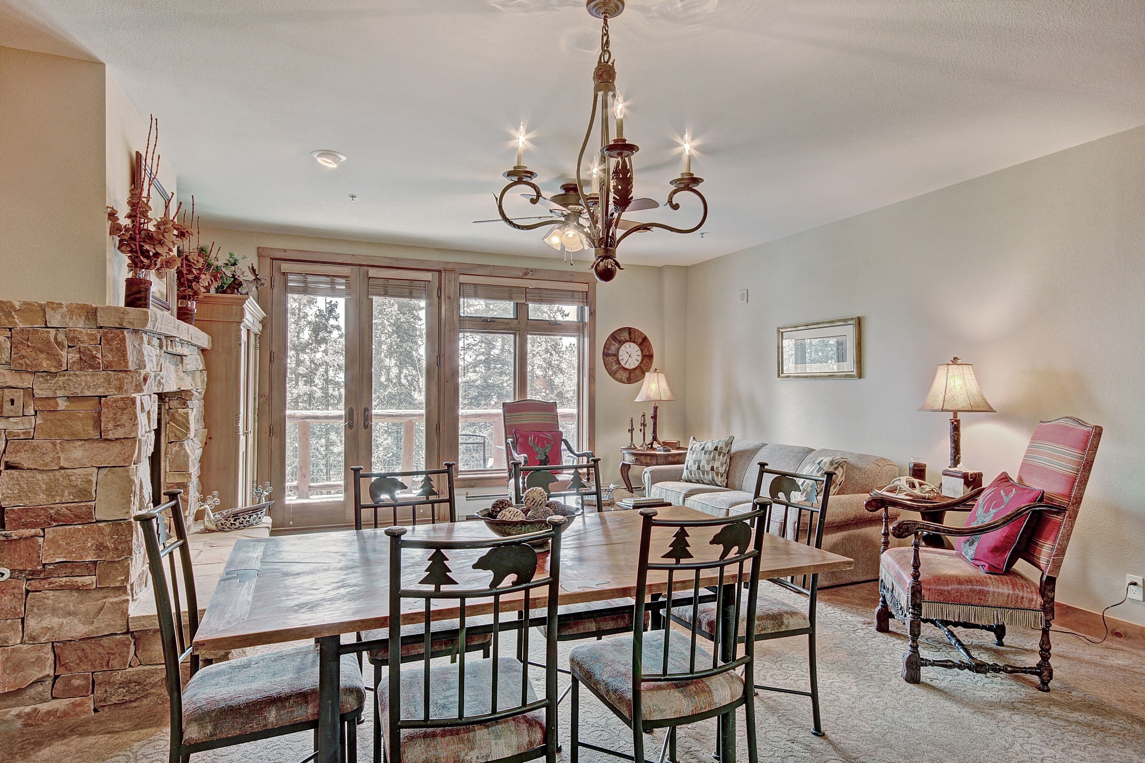 A dining area with a stone fireplace, a chandelier, and a large window with a view of trees.