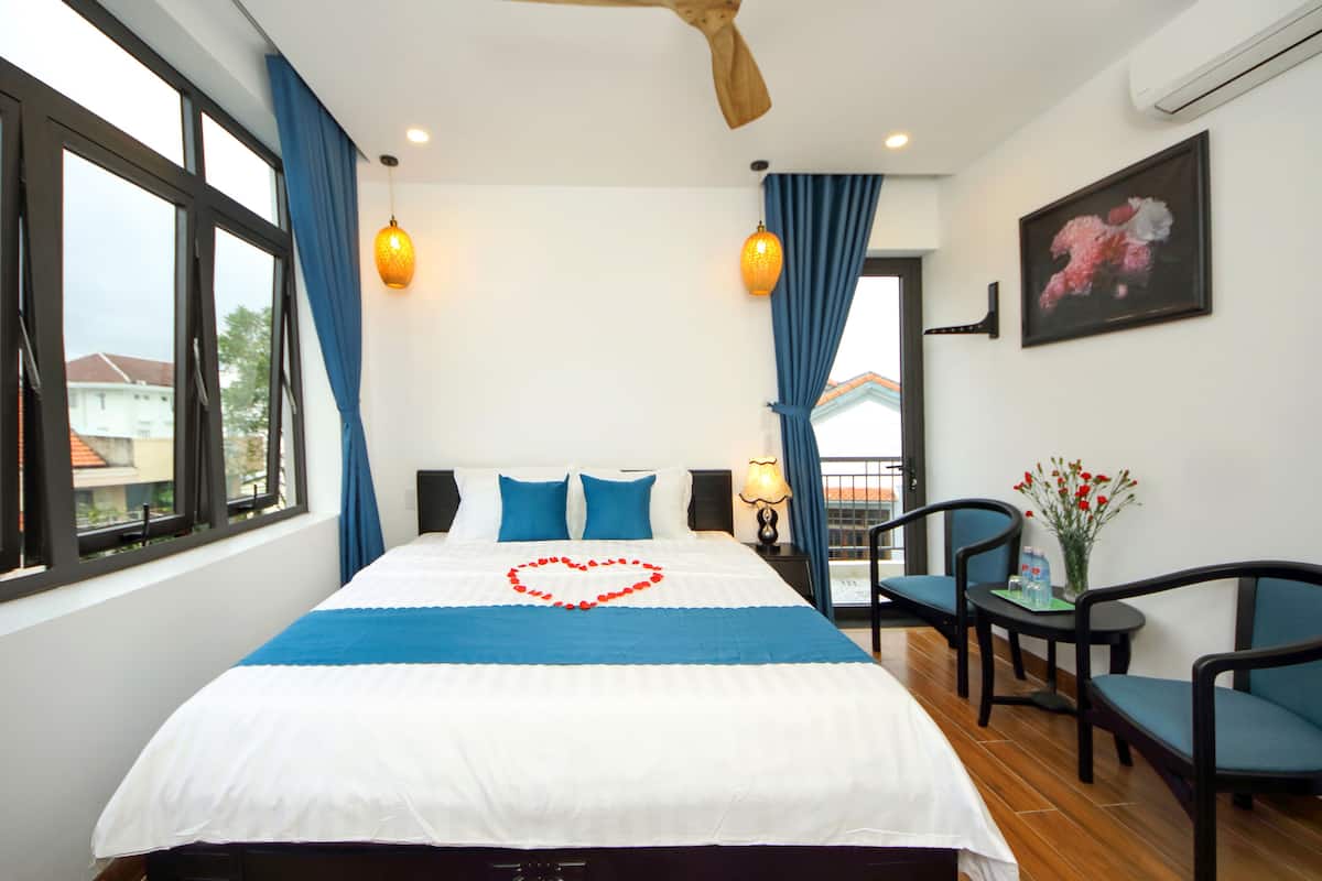 A hotel room with a large bed, a ceiling fan, a view of the city, and a small table with chairs.