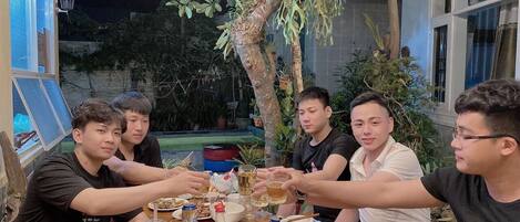 A group of people sitting around a table with food and drinks, in an outdoor setting with a tree and a building in the background.