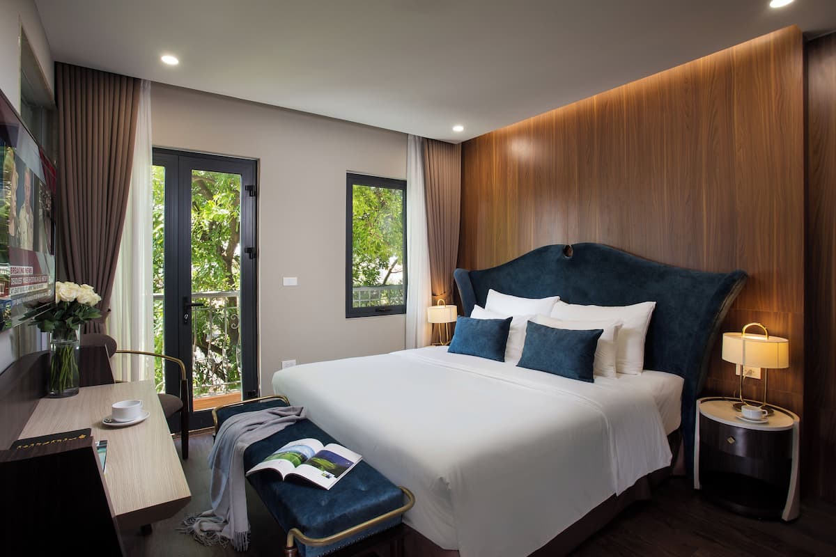 A modern hotel room with a large bed, a desk with a chair, a small table with a vase of flowers, and a bench.