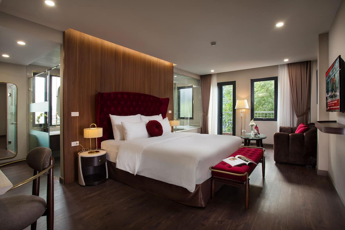 A modern hotel room with a large bed, a bedside table with a lamp, a small round table with a lamp, a chair, a sofa, and a large mirror.