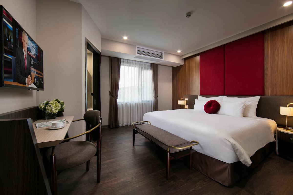 A hotel room with a large bed, a desk with a chair, a bench, a TV, and a window with curtains.