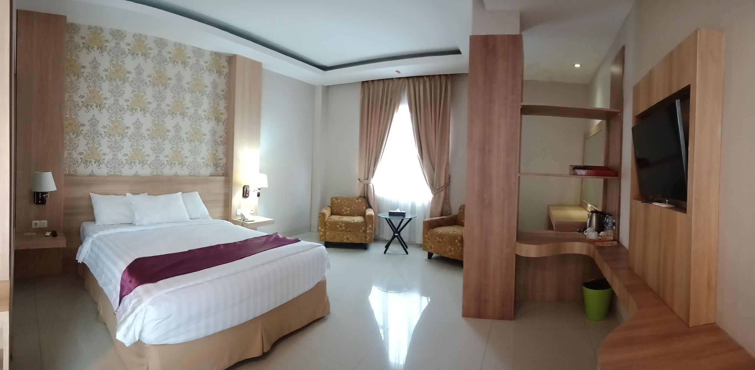A hotel room with a large bed, two armchairs, a small table, a TV, and a wooden partition.