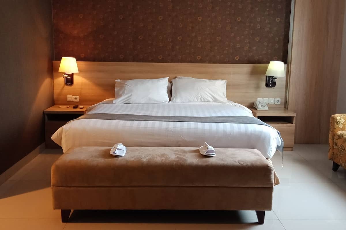 Ett hotellrum med en stor säng, två sängbord med lampor, en liten soffa och en pall med handdukar.
