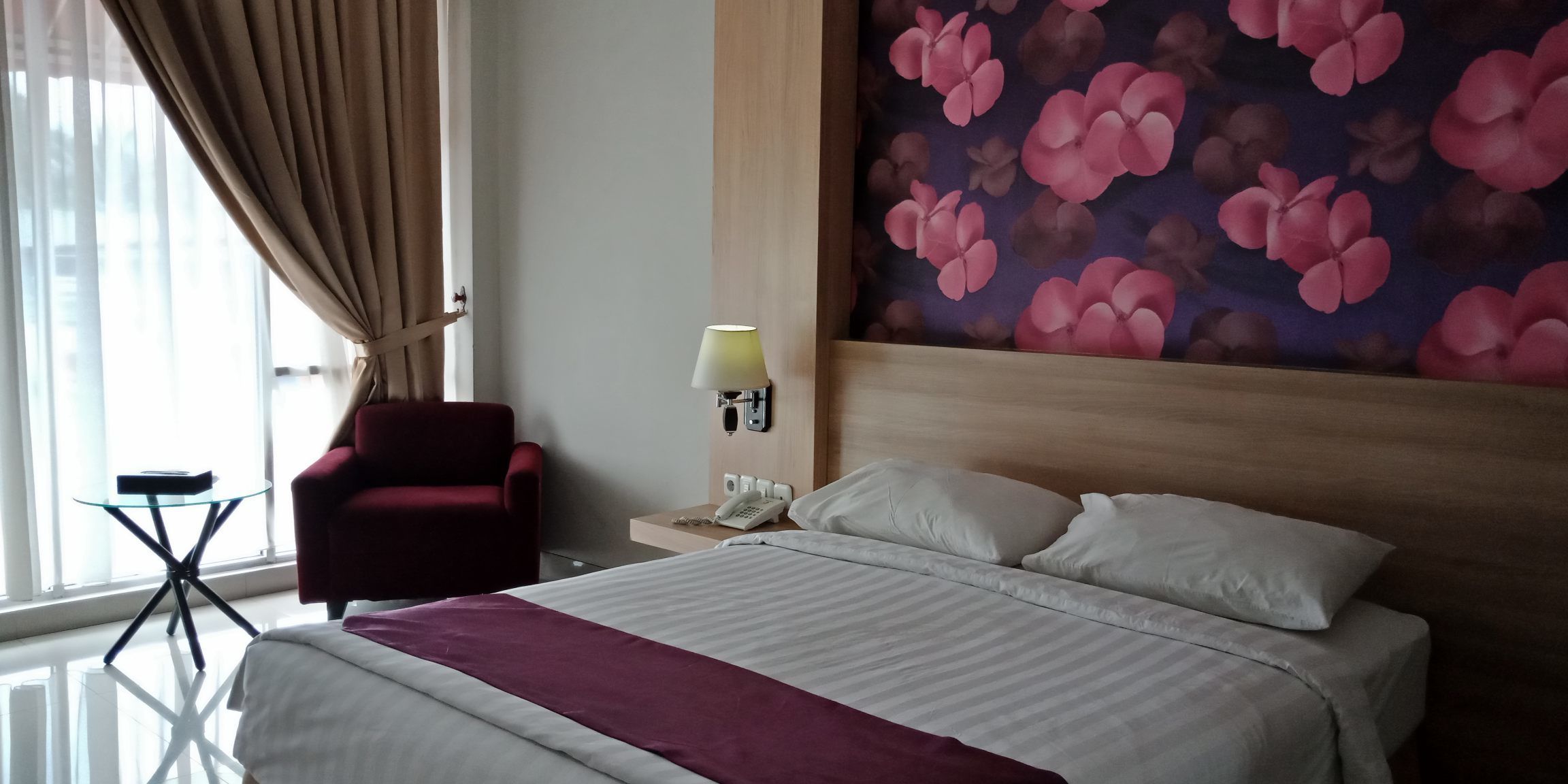 Habitación de hotel con una cama, una silla roja, una mesita, una lámpara y una pared con estampado floral.
