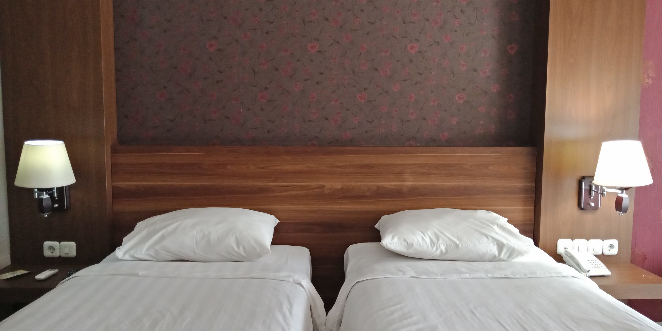 Une chambre d’hôtel avec deux lits, des têtes de lit en bois, un papier peint à motifs floraux et des lampes de chevet.