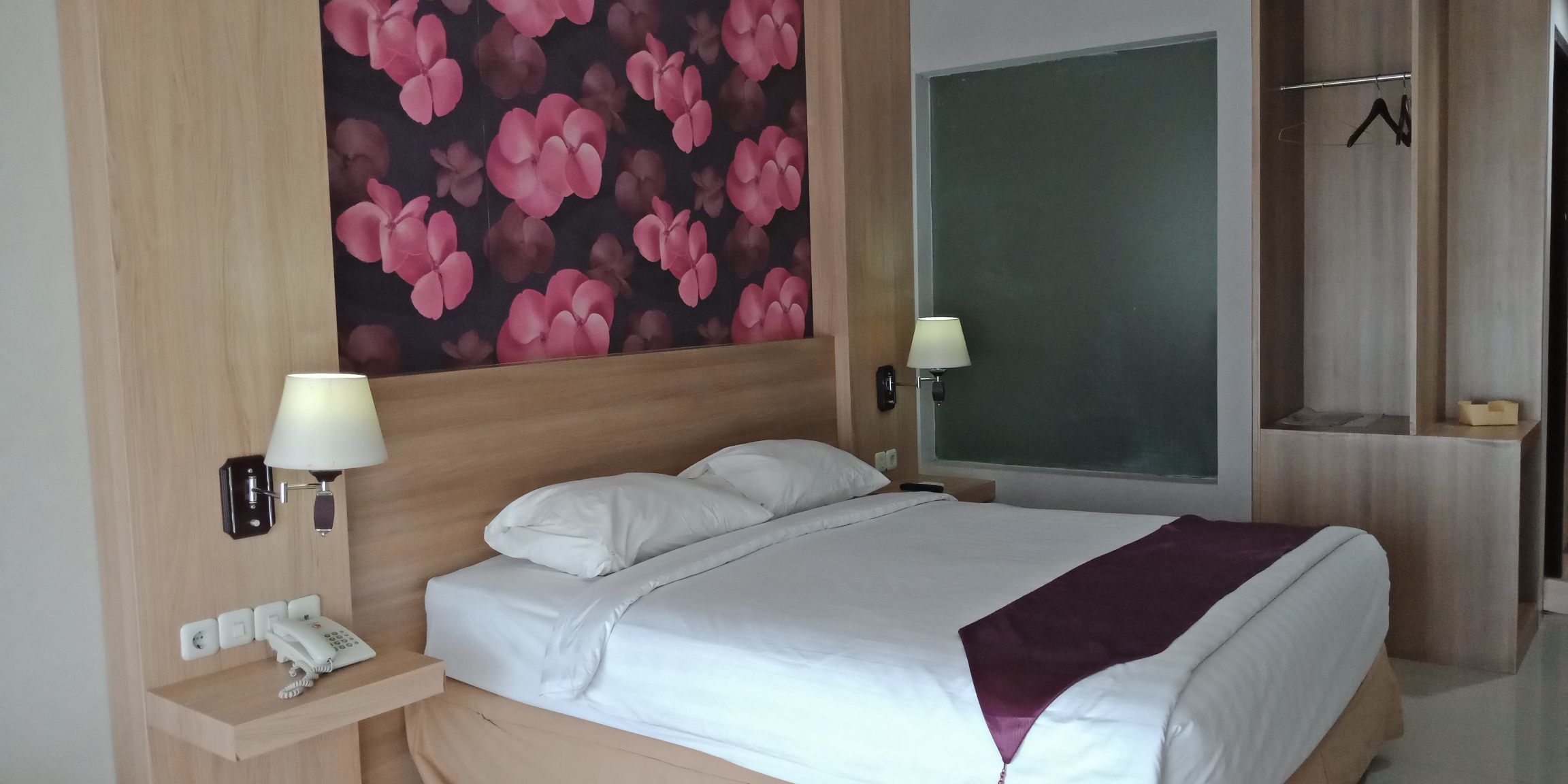 A hotel room with a large bed, two bedside lamps, a telephone, a wooden headboard, a wall with a floral pattern, and a closet with a hanger.