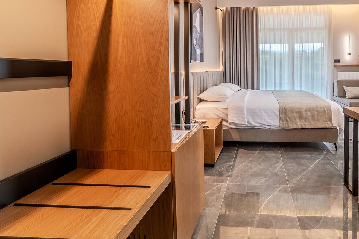 A modern hotel room with a wooden desk, a bed with a beige bedspread, a nightstand, and a large window with curtains.