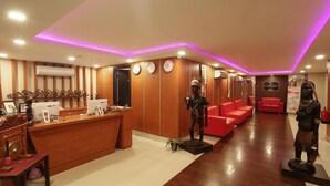 Un espace d’accueil d’hôtel avec un comptoir de réception en bois, des sièges rouges et des statues décoratives.