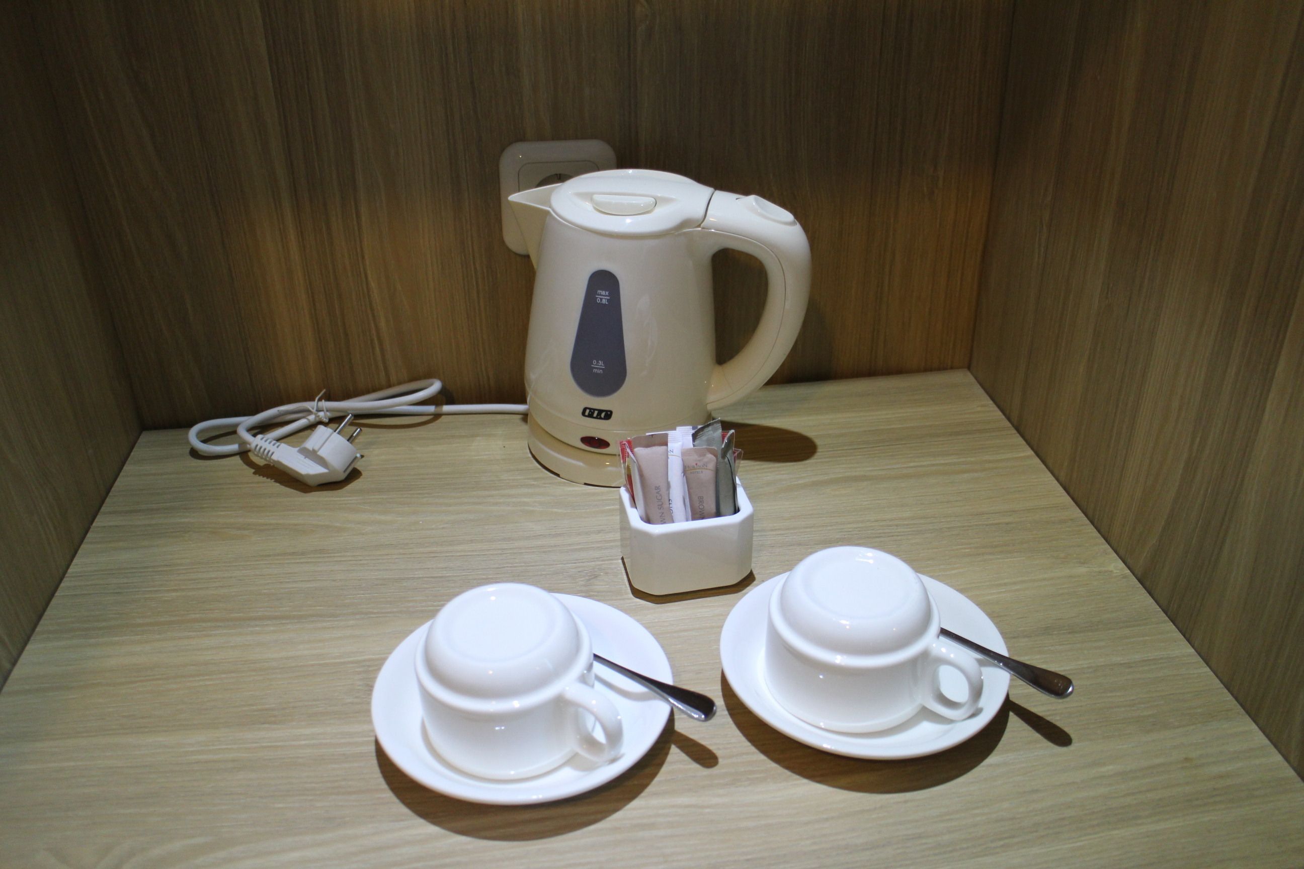 A wooden table with a white electric kettle, two white cups with saucers, and a small container holding a spoon and a packet of tissues.