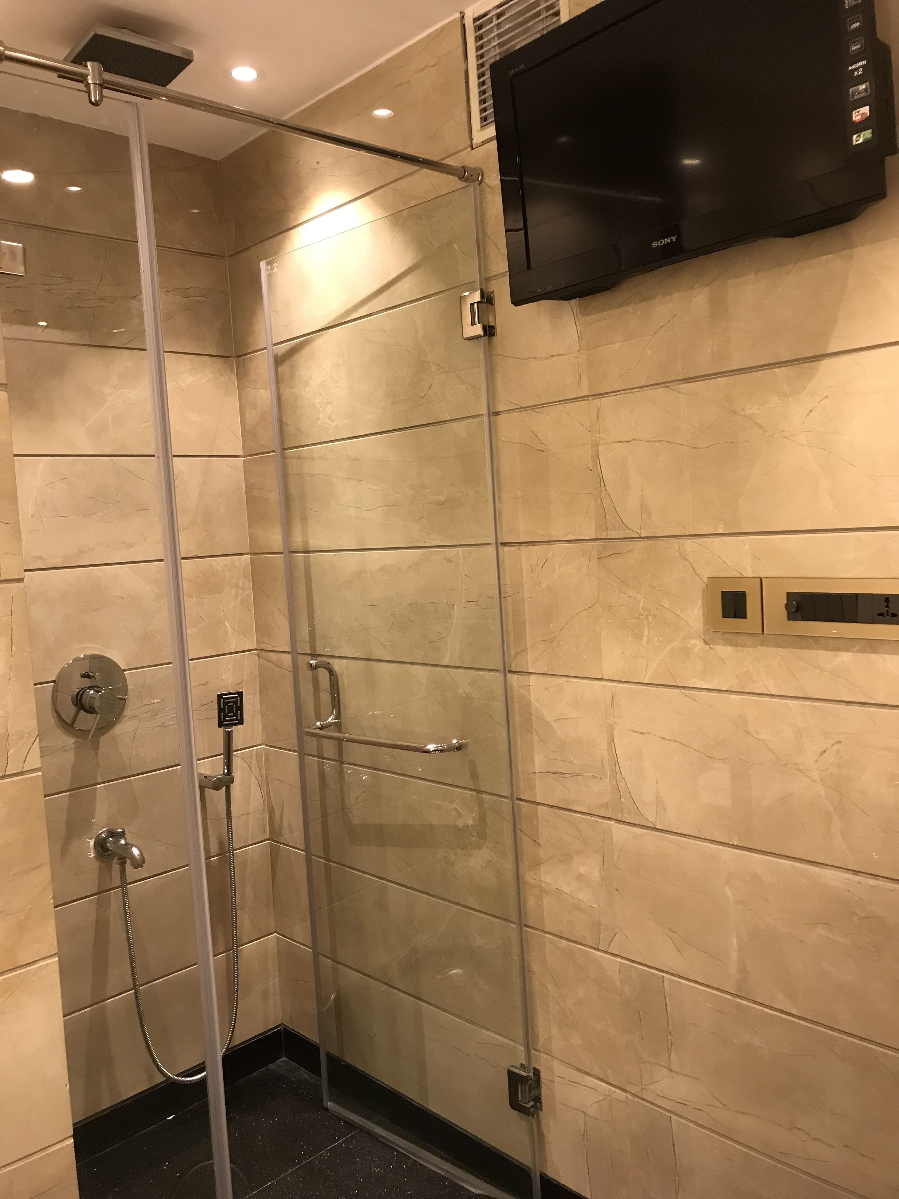 A modern bathroom with a glass-enclosed shower, a handheld showerhead, a wall-mounted TV, and a control panel.