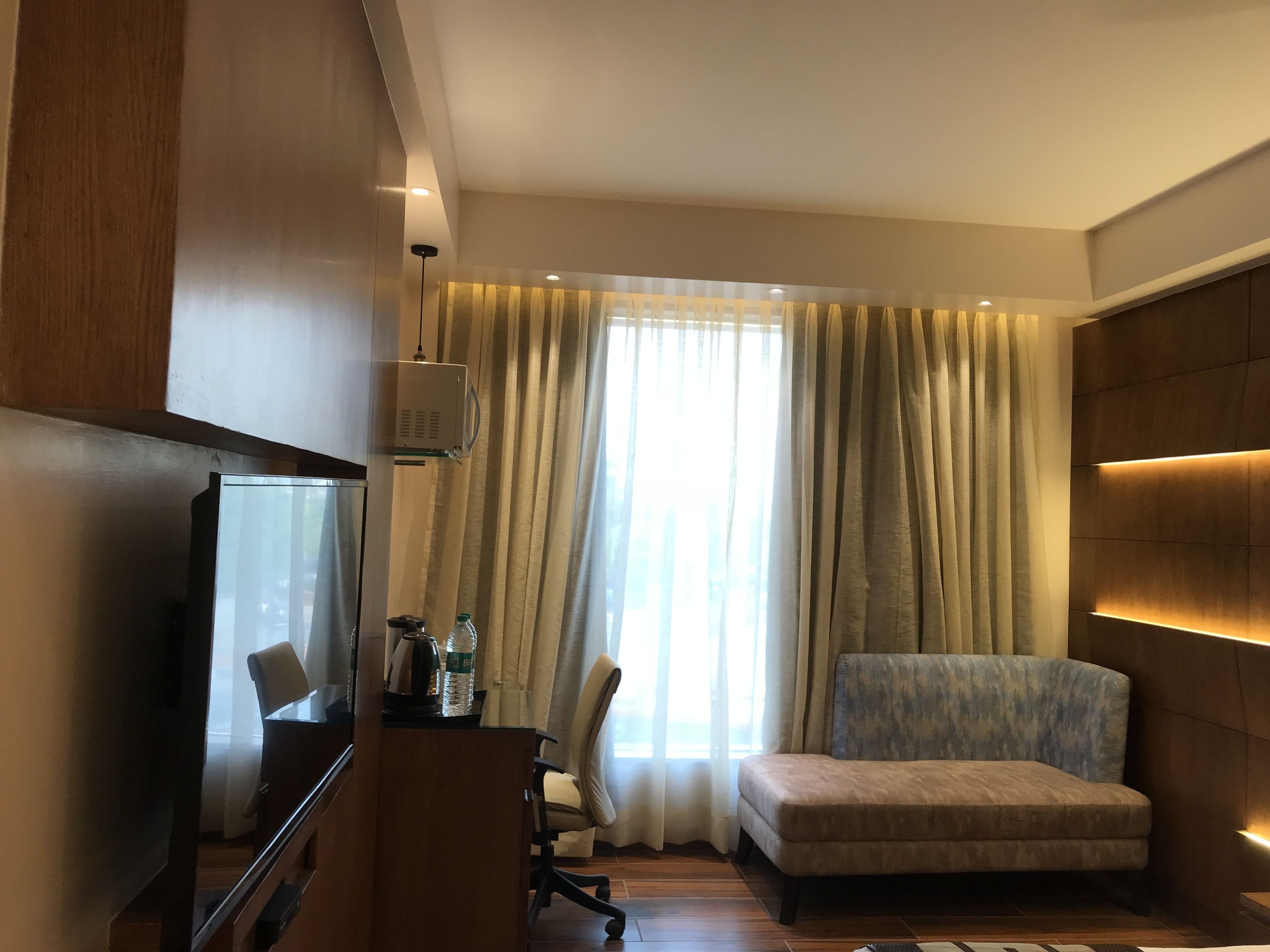 A modern hotel room with a flat-screen TV, a sofa, a desk with a lamp, and a large window with curtains.