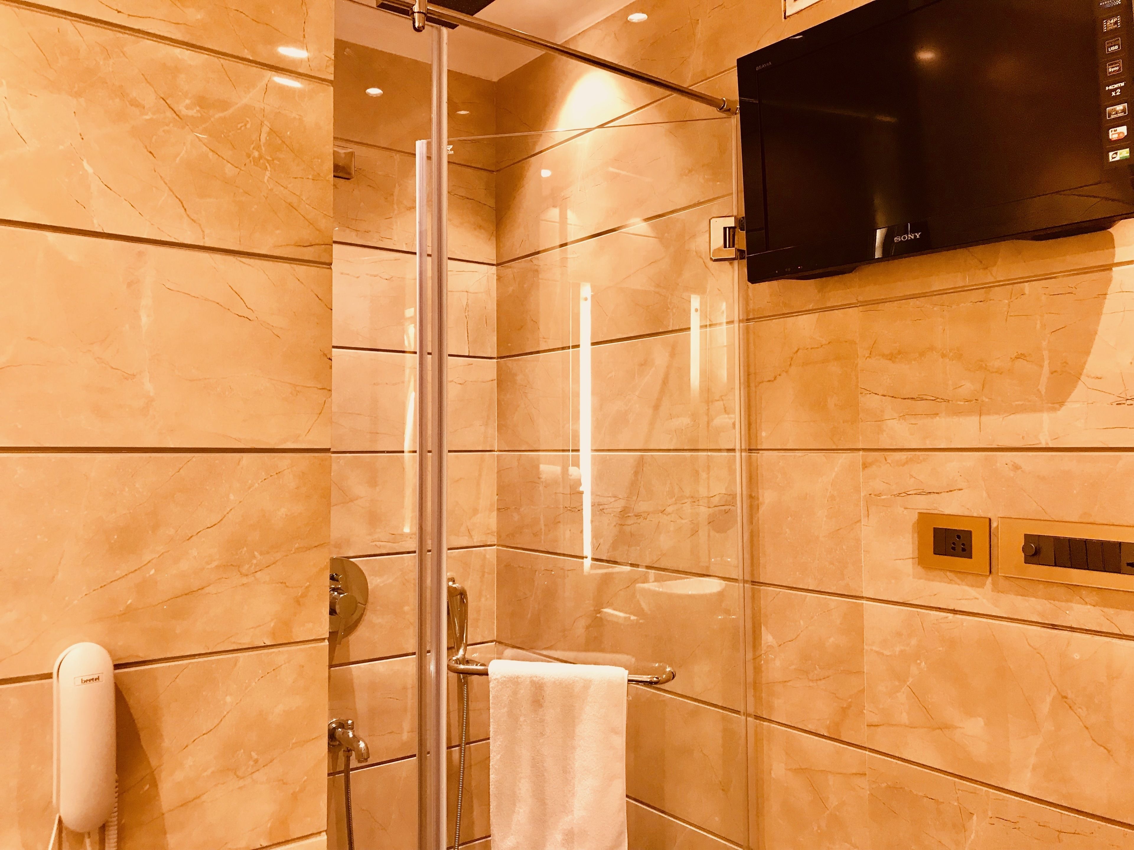 Baño con ducha envuelta en vidrio, televisor montado y dispensador de papel higiénico.