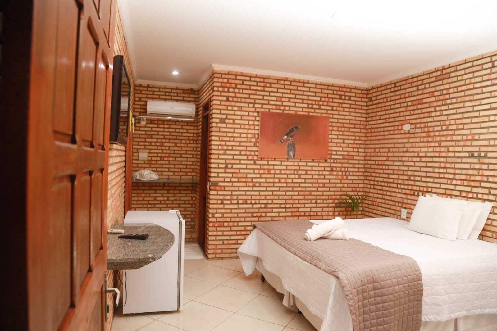 A hotel room with a brick wall, a bed with white bedding, a small desk with a towel, and a picture of a bird on the wall.