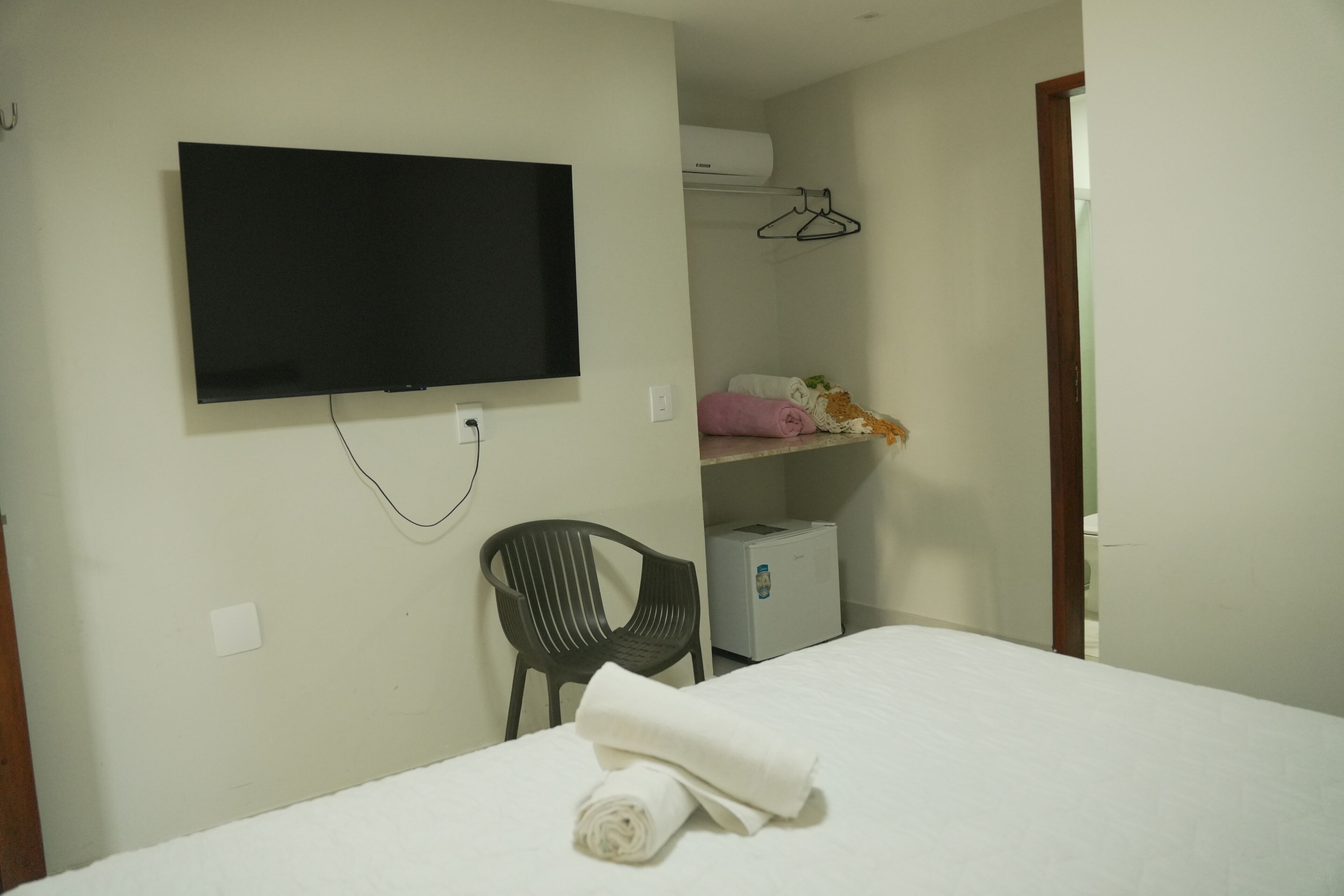 A hotel room with a bed, a TV mounted on the wall, a chair, a heater, and a wardrobe with hanging clothes.
