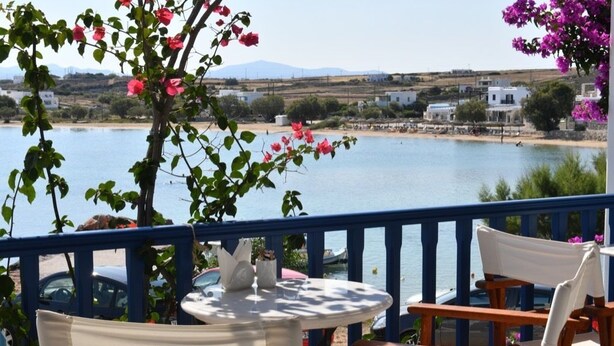 A terrace with a round table and chairs, overlooking a body of water with buildings in the background. - Bocamviglies By The Sea (Paros)