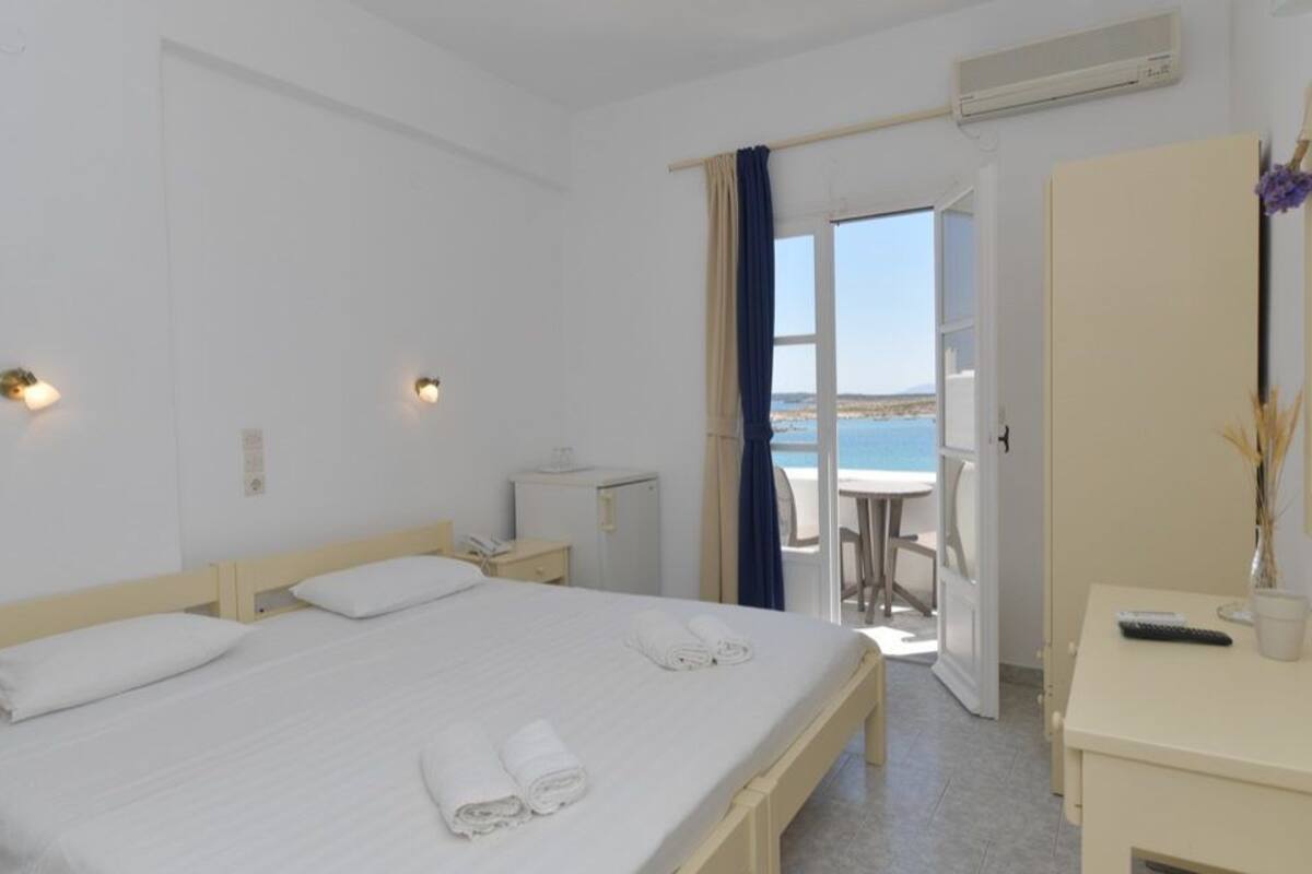 Una camera d'hotel con un letto, un comodino e una scrivania con una sedia. C'è una finestra con tende, un condizionatore d'aria e vista sul mare.