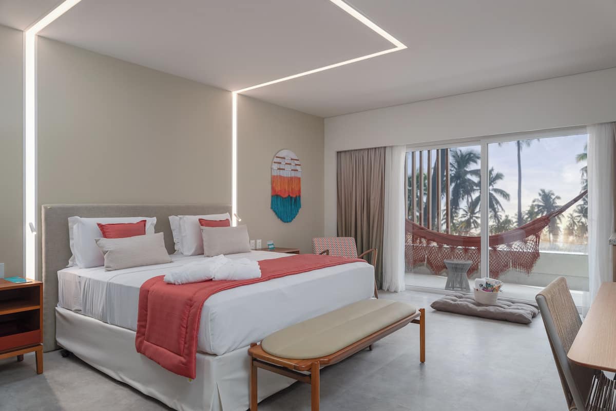 Une chambre d’hôtel moderne avec un grand lit, une banquette, une petite table et une vue sur les palmiers à travers de grandes fenêtres.