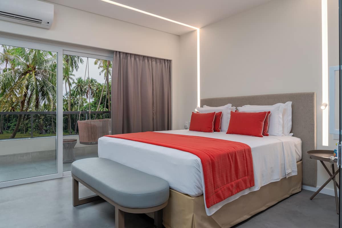 Une chambre d’hôtel moderne avec un grand lit, une banquette, une petite table et une vue sur les palmiers, visible à travers une porte-fenêtre coulissante.