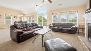Ein geräumiges Wohnzimmer mit Kamin, Ledersofas, einem Essbereich und großen Fenstern mit gestreifter Vorhangschiene.