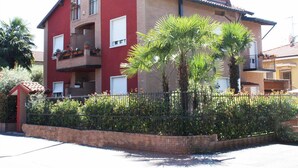 A two-story building with a red facade, balconies with potted plants, and a brick wall with greenery in front. - Mi Casa Affittacamere (Osio Sotto)